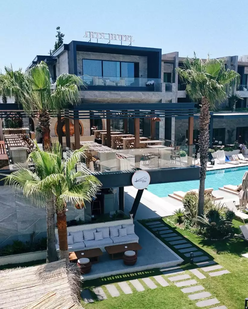 Bodrum'da yer alan ARTS HOTEL'in, üst katında balkonlu odalar, orta katında cam korkuluklu şık bir açık hava restoranı, alt katında ise SISHI yazılı yuvarlak tabela ile çevrili havuz kenarı ve çimli terasta konforlu oturma alanları bulunan modern ve lüks tesisinin genel görünümü.