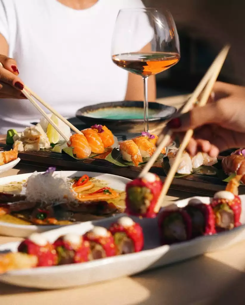 İki kişinin farklı türlerde somon nigiri, kırmızı rulolar ve diğer deniz ürünleri içeren özenle hazırlanmış suşileri çubuklarla alıp yediği, arka planda bir kadeh roze şarabın parladığı şık bir restoranda çekilmiş bu yakın plan görüntü, Bodrum'daki özel bir yemek deneyimini gözler önüne seriyor.