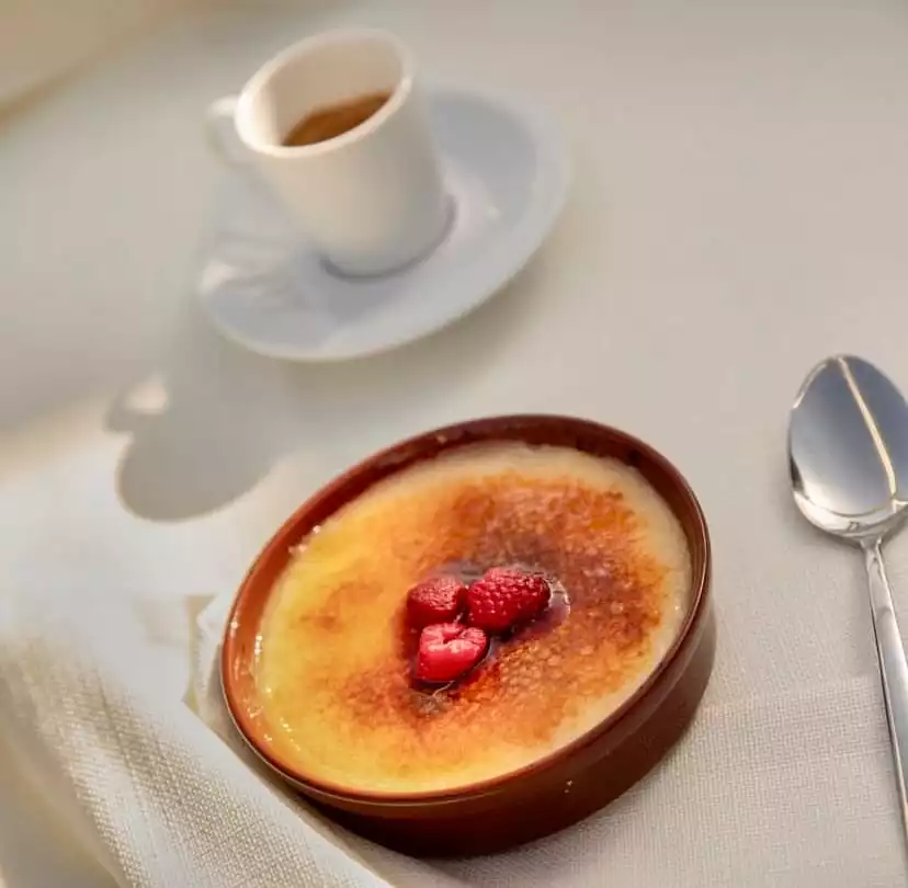 Üstü mükemmel karamelize edilmiş ve ortasına üç taze ahududu yerleştirilmiş, kahverengi bir kapta sunulan krem brulee tatlısının yakın çekiminde, arkada küçük bir fincan espresso ve yanında parlak bir metal kaşık açık renkli keten bir masa örtüsü üzerinde duruyor.
