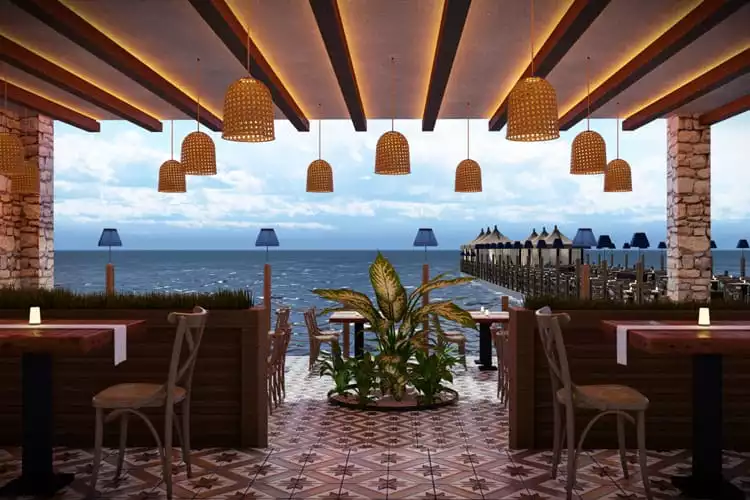 Deniz manzaralı bir restoranın içinden çekilmiş bu görselde, hasır avizelerle aydınlatılmış, ahşap masalar ve desenli yer karolarına sahip ferah bir oturma alanı ile uzakta iskele üzerindeki şemsiyelerin yarattığı Bodrum tatil atmosferi vurgulanıyor.