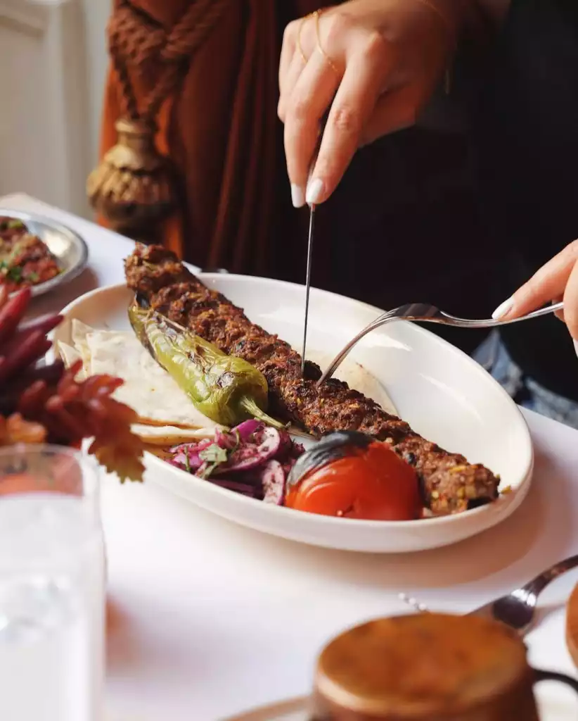 Beyaz oval bir tabağın üzerinde ızgara biber, közlenmiş domates ve sumaklı soğan salatası eşliğinde sunulan Adana veya Urfa kebabı, ince lavaş ekmeği üzerinde, bir misafirin bıçak ve çatalla özenle kestiği anı yansıtırken, Türk mutfağının lezzetlerini gözler önüne seriyor.