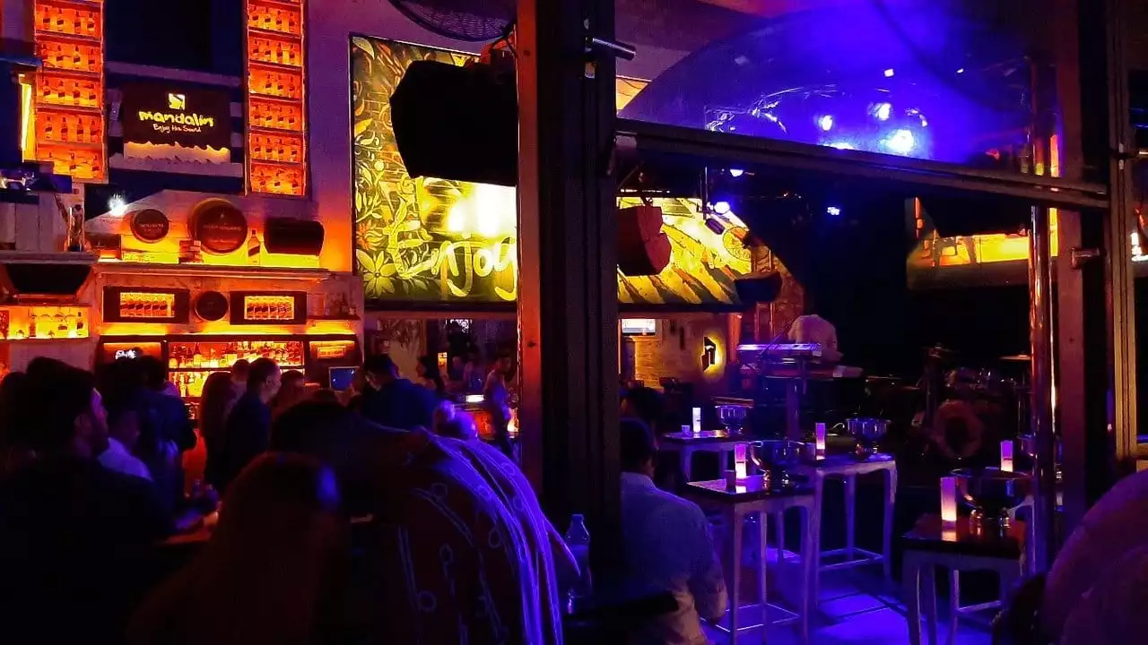 Bodrum'un hareketli gece hayatını yansıtan bu canlı mekanda, turuncu ışıklarla aydınlatılmış Mandalin barında içki şişeleri göz alırken, sahne kısmında ise mavi-mor ışıklar altında bir DJ performans sergiliyor ve insanlar keyifli anlar yaşıyor.