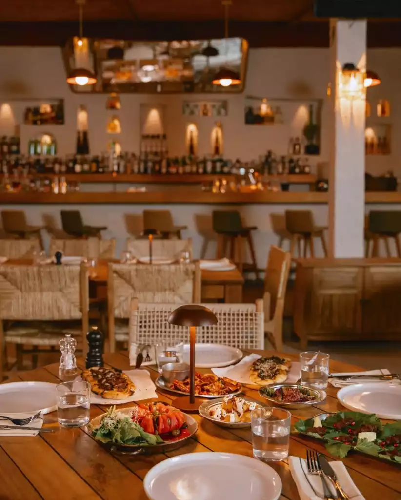 Bodrum'un seçkin bir restoranında, özenle hazırlanmış domatesli salata, nar taneli yeşillikler, çeşitli mezeler ve pidelere benzer lezzetlerle dolu ahşap bir masada şık bir yemek düzeni sergileniyor; arka planda ise modern bar alanı ve sıcak aydınlatma mekanı tamamlıyor.