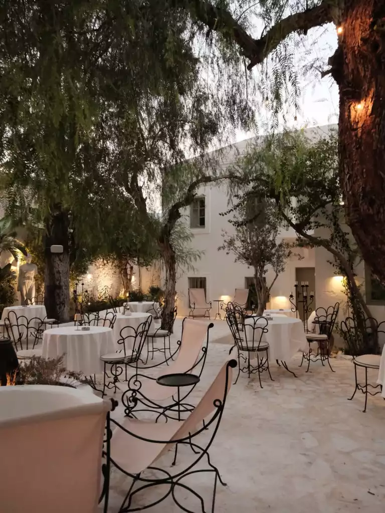Bodrum'a özgü bembeyaz bir avluda, ışıklarla aydınlatılmış ağaçların altında, zarif beyaz örtülü masalar ve konforlu ferforje sandalyelerle düzenlenmiş, sakin bir akşam yemeği veya keyifli sohbetler için eşsiz bir atmosfere sahip bir açık hava mekanı.