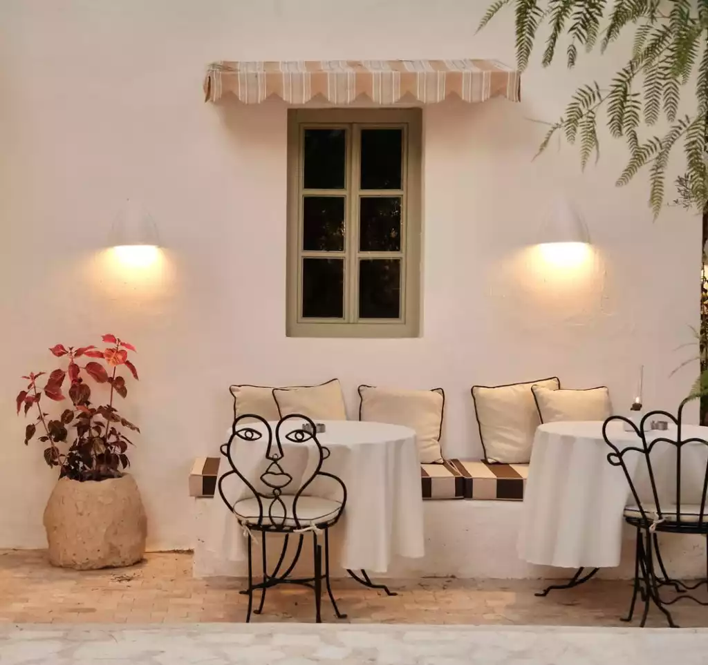 Bodrum'un beyaz badanalı duvarlarına yaslanmış, üzerinde çizgili tente ve ahşap görünümlü bir pencere bulunan, iki duvardan aydınlatmalı sıcak bir kafe köşesi. Ortamda sanatsal metal sırtlı sandalyeler, beyaz örtülü yuvarlak masalar, arkada çizgili minderli ve yastıklı bir bank ile köşede kırmızı yapraklı bir bitki, davetkar ve sakin bir atmosfer sunuyor.