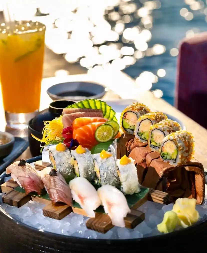 Bodrum'da deniz kenarında, parıldayan güneşli su manzarası eşliğinde sunulan, somon ve ton balığı sashimi, çeşitli maki ve nigiri çeşitlerini içeren, buz üzerinde özenle hazırlanmış bir sushi tabağı ve yanında ferahlatıcı portakal kokteyli.