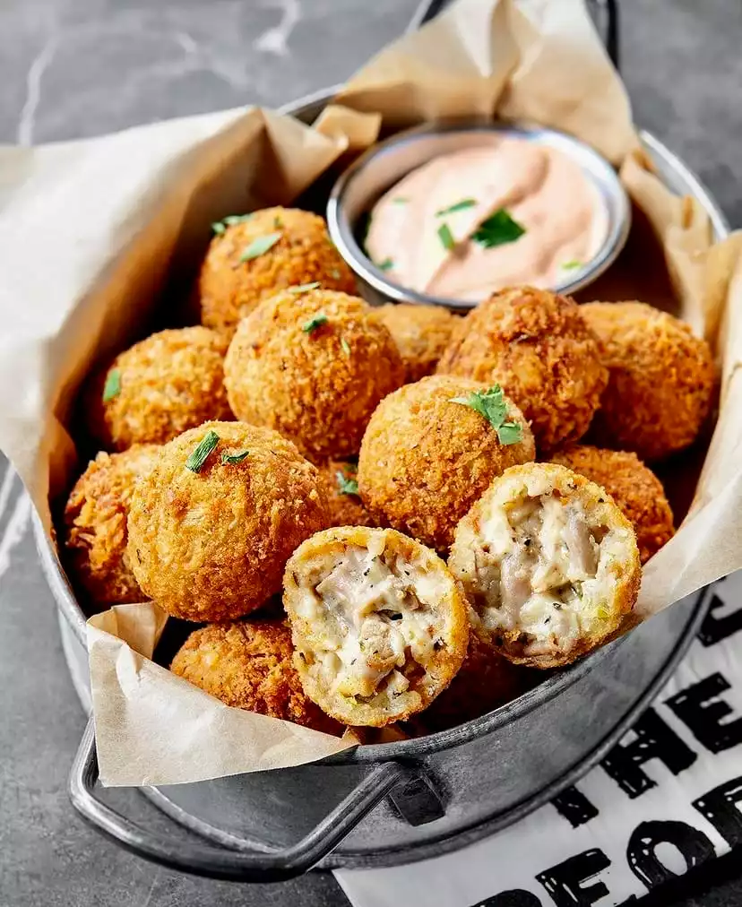 İçi kremsi peynir ve mantar dolgulu, dışı çıtır altın rengi paneli arancini benzeri kızarmış toplar, yanlarında pembe renkli bir sos eşliğinde, rustik metal bir kap içinde yağlı kağıt üzerinde iştah açıcı bir şekilde sunuluyor.