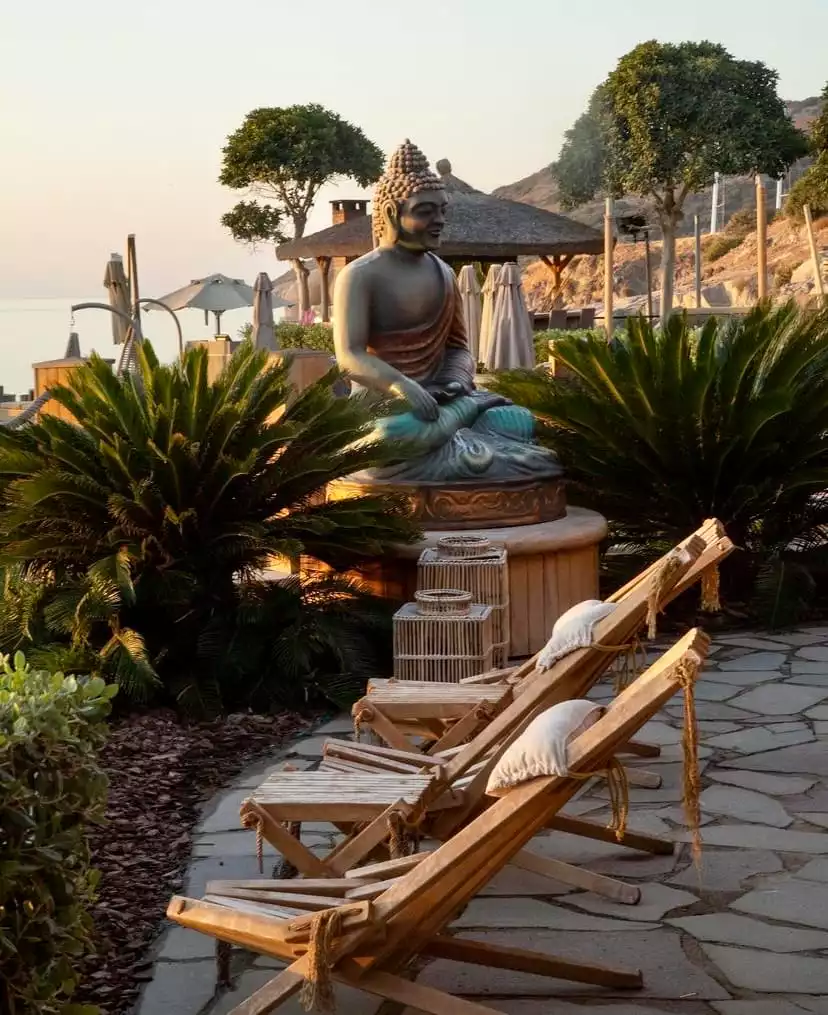 Bodrum'un en iyi restoranlarından biri olan Buddha Bar'ın bulunduğu huzurlu bir koyda, altın rengi gün batımı ışıkları altında oturmuş büyük bir Buda heykeli, çevresindeki tropikal bitkiler ve ön plandaki şık ahşap şezlonglarla birlikte sakin bir Akdeniz ambiyansı sunuyor.