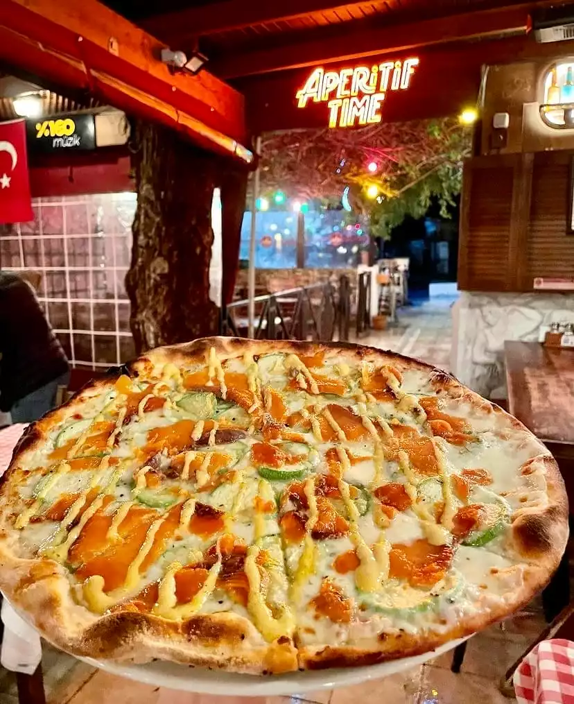 Ortada, füme somon, kabak dilimleri ve kremalı soslarla zenginleştirilmiş büyük, lezzetli bir pizza, arkada akşam ışıklarında parlayan Aperitif Time neon tabelası ve hareketli bir Bodrum restoranı ortamı görülüyor.