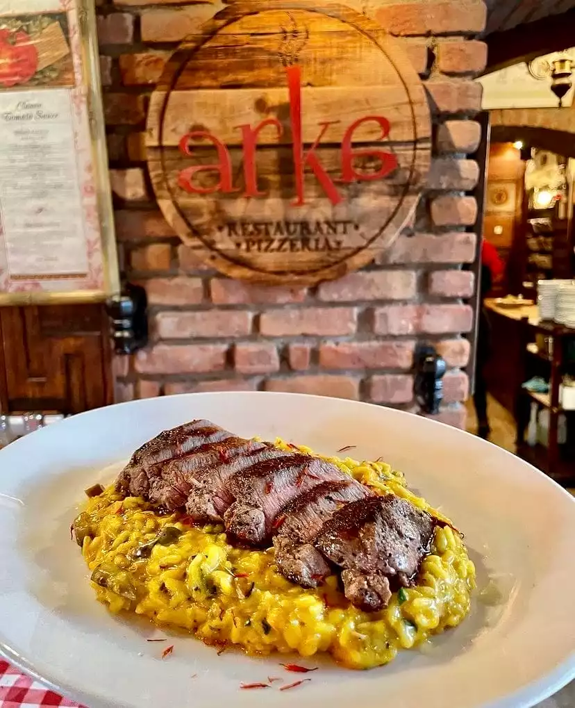 Bodrum'daki Arke Restaurant Pizzeria'da sunulan bu çekici yemekte, parlak sarı renkli risotto üzerinde ustaca ızgaralanmış ve dilimlenmiş dana eti yer alırken, arka planda tuğla duvar ve Arke Restaurant'ın rustik ahşap tabelası görülüyor.