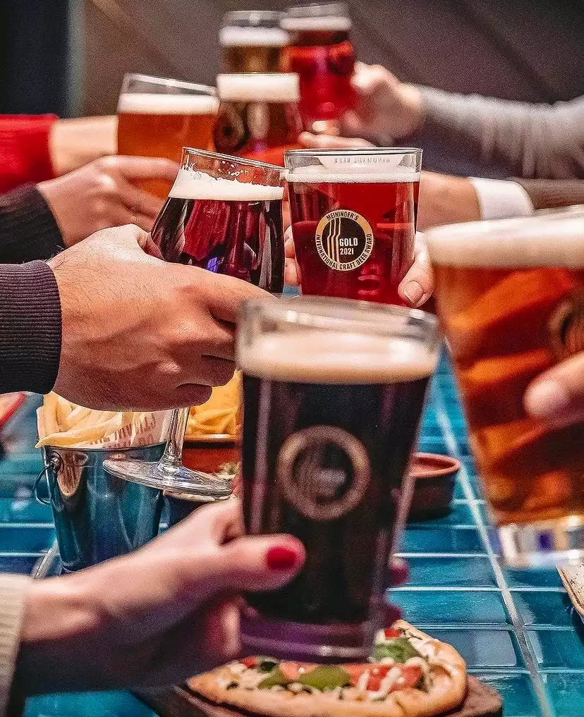 Birden fazla kişinin ellerinde farklı renklerde bira bardaklarını kadeh kaldırarak tuttuğu, ortamda bira ödülü etiketi görünen bir zanaat birası ile pizza ve patates kızartması eşliğinde keyifli bir sosyal buluşma anı.