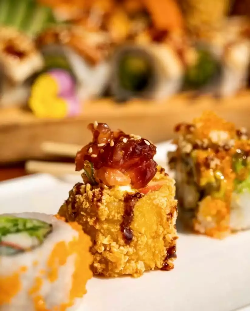 Bodrum'un gastronomi sahnesinden bir kesit sunan bu karede, beyaz bir tabakta sunulan, üzeri çıtır kızarmış pirinçle kaplı, parlak koyu renkli bir sos ve bol susam ile süslenmiş, üzerinde nar tanelerine benzeyen kırmızımsı bir garnitür bulunan gösterişli bir sushi rulosu ön planda tüm cazibesiyle dururken, arka planda farklı çeşitlerdeki diğer lezzetli sushiler bulanık bir şekilde masadaki zengin sunumu tamamlıyor.