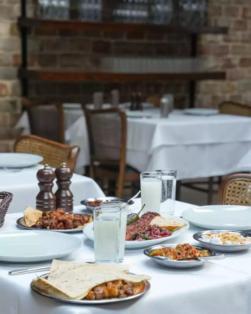 Beyaz masa örtülü bir restoranda, şiş kebap, kuşbaşı et, lavaş ekmeği gibi ana yemeklerin yanı sıra çeşitli mezeler ve ayran bardaklarıyla donatılmış, misafirlerini bekleyen iştah açıcı bir Türk sofrası, loş ışıklı tuğla duvarlı ve ahşap raflı otantik bir mekanda sıcak bir atmosfer sunuyor.