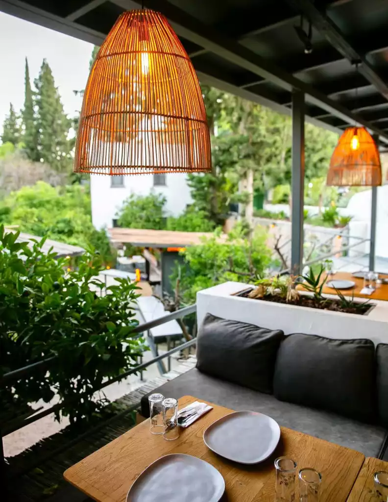 Bodrum'da bulunan modern bir açık hava restoranın samimi atmosferini yansıtan bu geniş teras, parlak bambu avizelerin ışığı altında, hazırlanmış ahşap masalarda boş tabaklar ve bardaklar ile konuklarını bekliyor gibi duruyor, arka planda ise yemyeşil bitki örtüsü ve ağaçlarla çevrili huzurlu bir manzara uzanıyor.