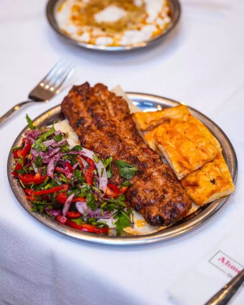 Nar gibi kızarmış, uzun bir kebap, yanında taze kırmızı soğan, biber ve yeşilliklerle hazırlanmış bol salata ile sıcak pide dilimlerinin bembeyaz bir masa örtüsü üzerinde metal bir tabakta servis edildiği, arka planda bulanık bir şekilde yoğurtlu bir mezenin de göründüğü iştah açıcı bir yemek tabağı.