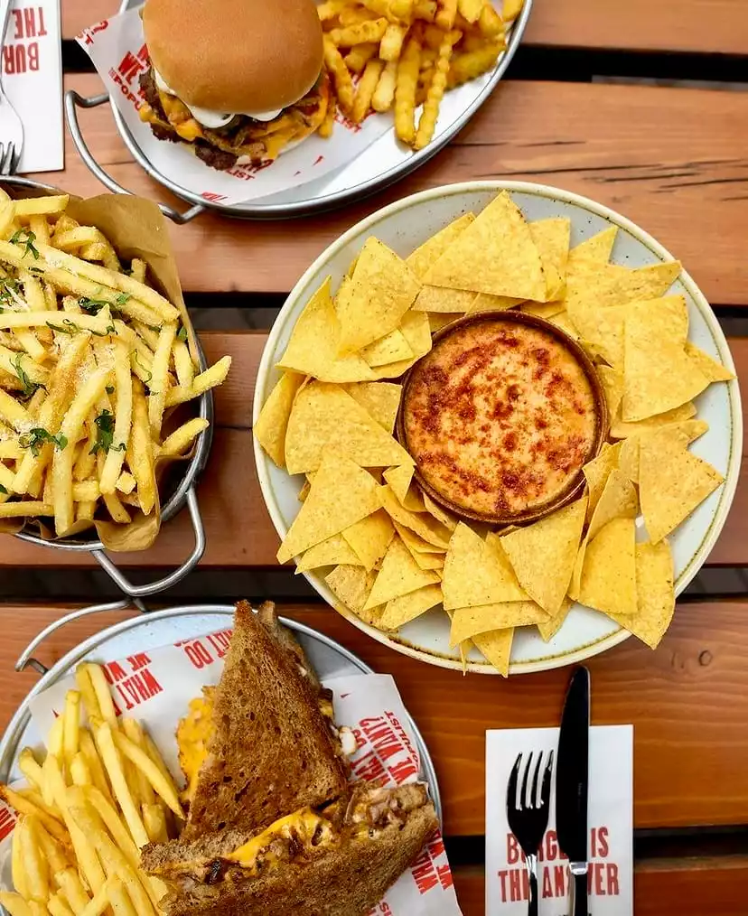 Ahşap bir masanın üstten çekilen bu zengin fotoğrafında, bol patates kızartması eşliğinde iştah açıcı bir çift köfteli burger, eritilmiş peynir soslu üçgen nacho cipsleri ve erimiş peynirle dolu bir ızgara sandviç, hepsi metal servis tepsileri ve kaselerde sunularak keyifli bir yemek ziyafeti vaat ediyor.