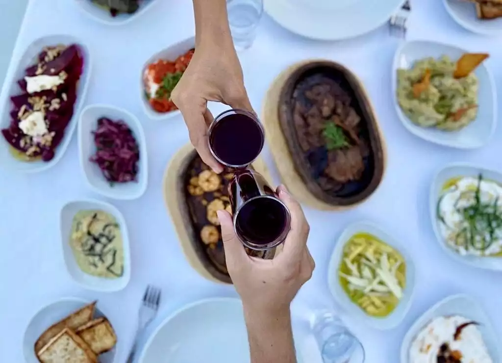 Bodrum'un zengin mutfağını yansıtan beyaz örtülü bir masada, cevizli pancar salatası, kırmızı lahana turşusu, sarımsaklı yoğurtlu mezeler, karides tava ve güveçte et gibi birçok küçük kasede sunulan lezzetli çeşitlerin ortasında iki el, koyu renkli içecek dolu kadehleri neşeyle tokuşturarak keyifli bir yemeğin tadını çıkarıyor.