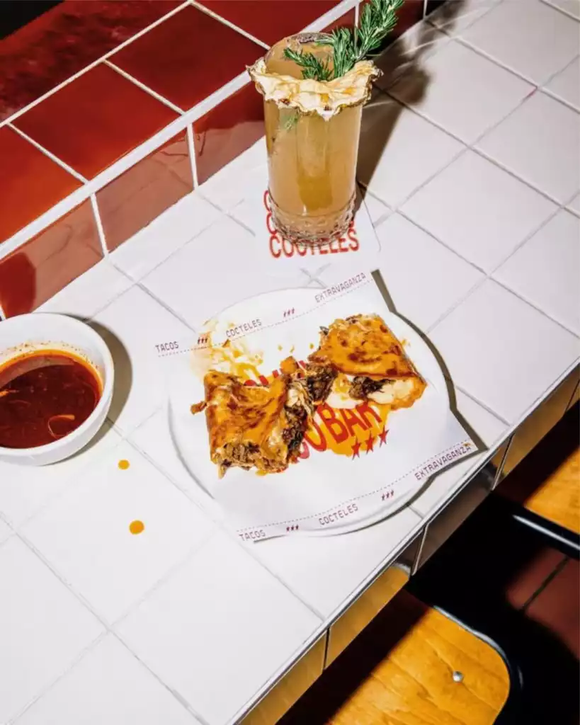 Beyaz karolu bir tezgah üzerinde, üzerinde TACOS COCTELES EXTRAVAGANZA yazılı kağıtla servis edilmiş iki adet kızarmış taco, yanında küçük bir kase kırmızı sos ve biberiye ile süslenmiş, ananas dilimli amber renkli bir kokteyl, Bodrum'un enerjik yeme içme kültürünü yansıtan modern bir bar ambiyansını sergiliyor.
