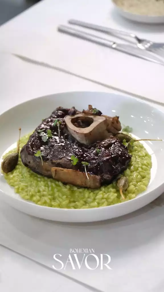 Beyaz bir masa üzerinde, kemikli etin parlak koyu sosu ve taze yeşil filizlerle bezenmiş, kremalı ve canlı yeşil risotto yatağında zarifçe sunulduğu bu sanatsal tabak, Bodrum'un kaliteli restoranlarında keşfedilebilecek özel lezzetleri ve sofistike bir yemek deneyimini gözler önüne seriyor.