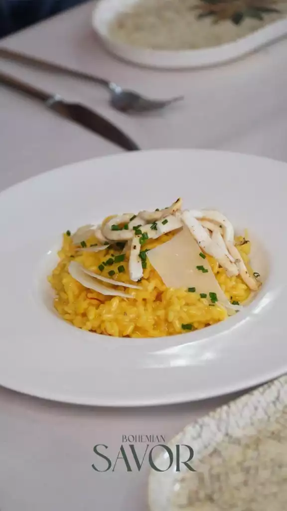 Ortada duran beyaz tabağın içinde safranlı risotto, üzerinde ızgara kalamar, rendelenmiş parmesan peyniri ve doğranmış taze soğanla süslenmiş, arka planda bulanık bir masada çatal bıçak ve başka bir yemekle Bodrum'un lezzetlerini yansıtan iştah açıcı bir sunum.