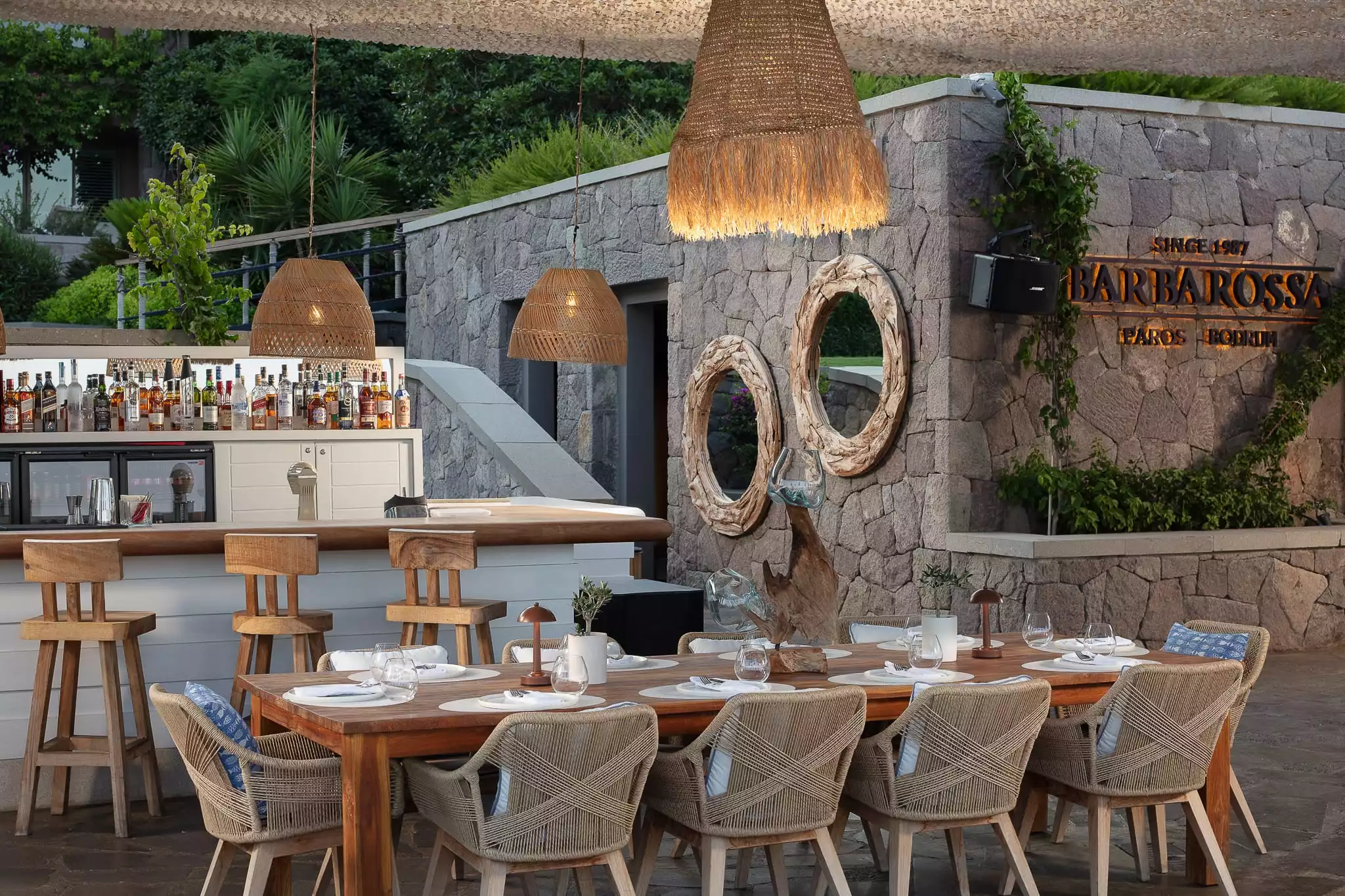 Bodrum'daki Barbarossa restoranının açık hava mekanında, büyük bir ahşap yemek masası etrafında örgü sandalyeler, şişelerle dolu bir bar, dekoratif örgü sarkıt lambalar ve Barbarossa tabelalı rustik taş duvarlar, yemyeşil bitkilerle çevrili sıcak ve davetkar bir ambiyans yaratıyor.