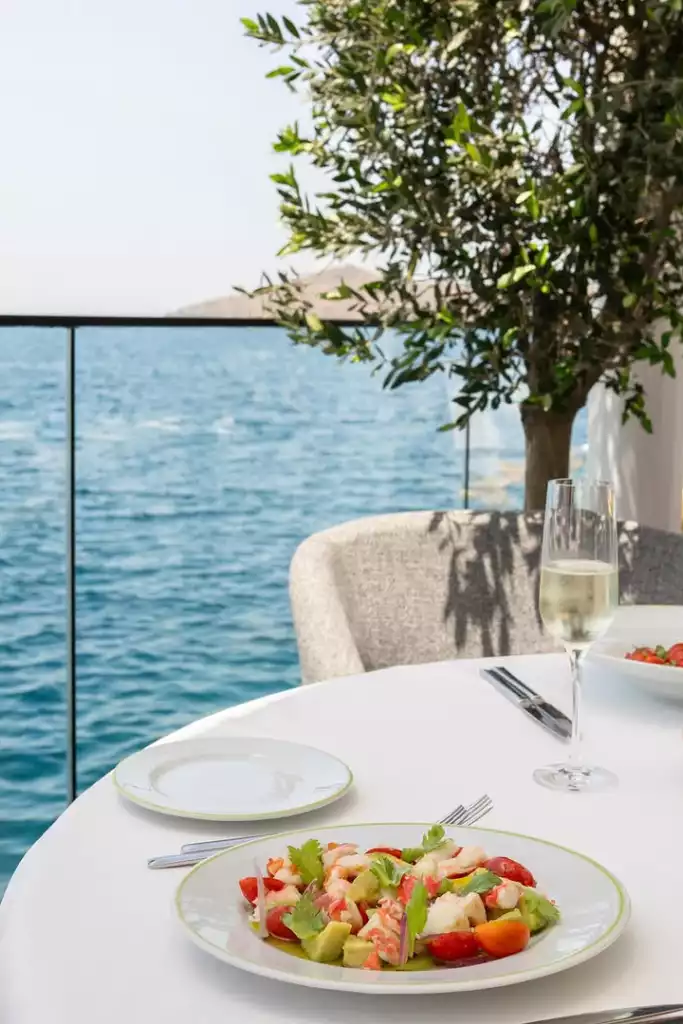 Bodrum'daki şık bir açık hava restoranında, masmavi Ege Denizi'nin büyüleyici manzarasına karşı, zeytin ağacının hoş gölgesinde yer alan beyaz yuvarlak bir masada taze karides, avokado ve çeri domatesli renkli bir salata ile zarif bir kadeh köpüklü şarap bulunmaktadır.