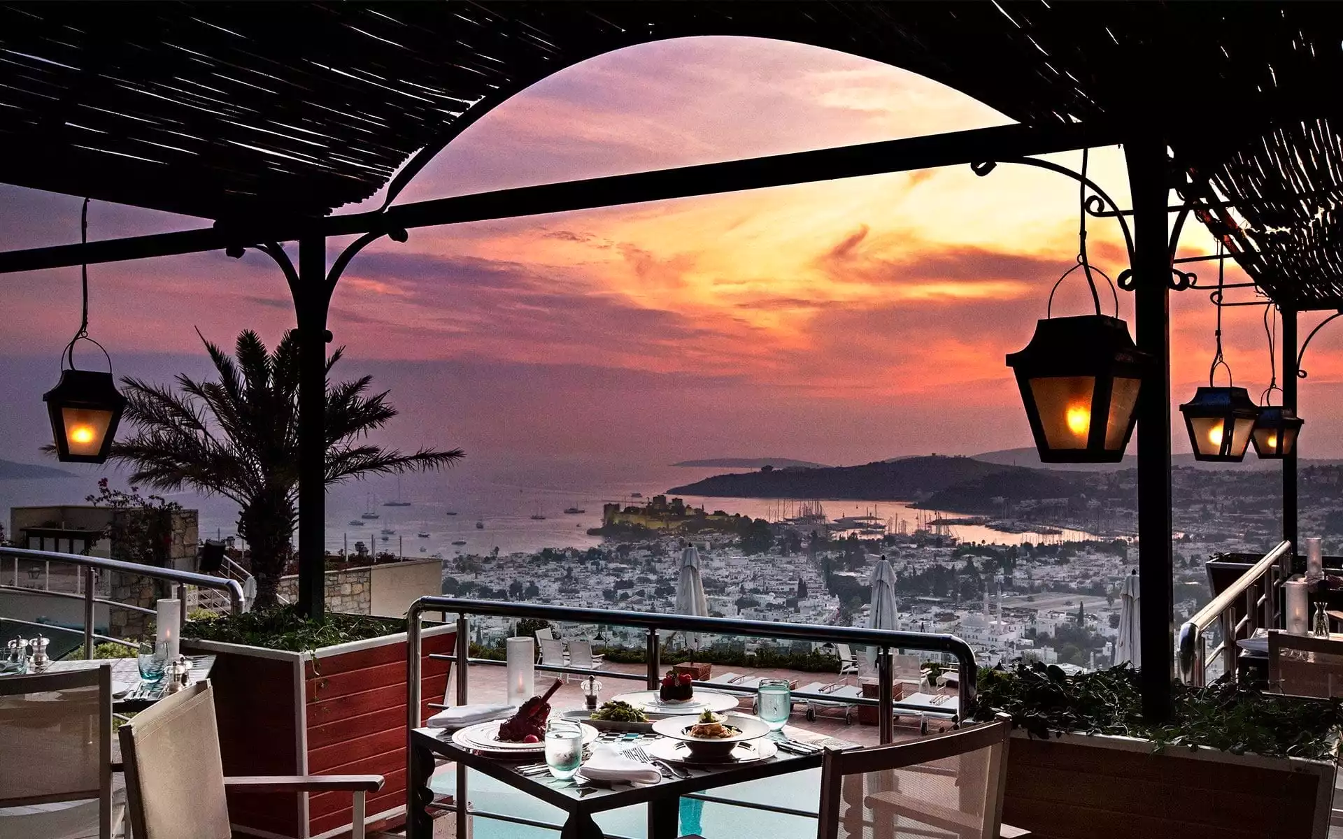 Gün batımının turuncu, pembe ve mor tonlarıyla aydınlanan gökyüzünün altında, Bodrum Kalesi ve teknelerle dolu denizin göz kamaştırıcı manzarasına sahip bir terasta, özenle hazırlanmış bir akşam yemeği masası romantik bir atmosfer sunuyor.