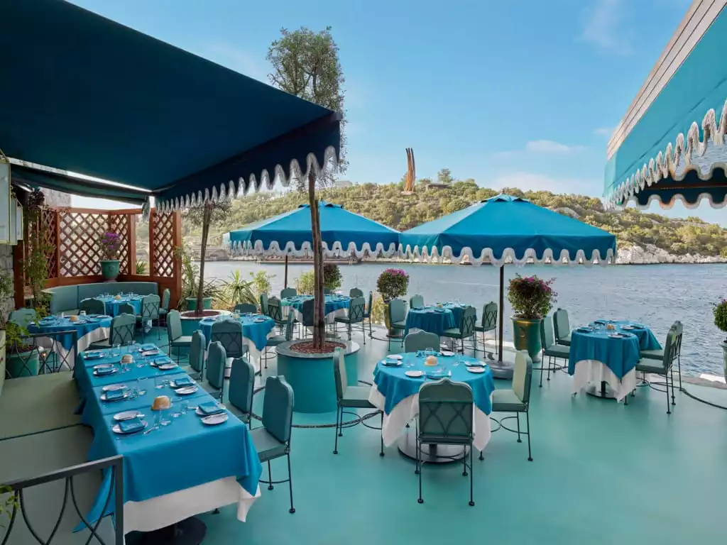 Bodrum'da denize sıfır konumdaki bu lüks restoranda, açık mavi masa örtüleri, zarif sandalyeler ve gölgelik sağlayan büyük şemsiyelerle donatılmış masalar, huzurlu bir yemek deneyimi için eşsiz bir Ege manzarası sunuyor.