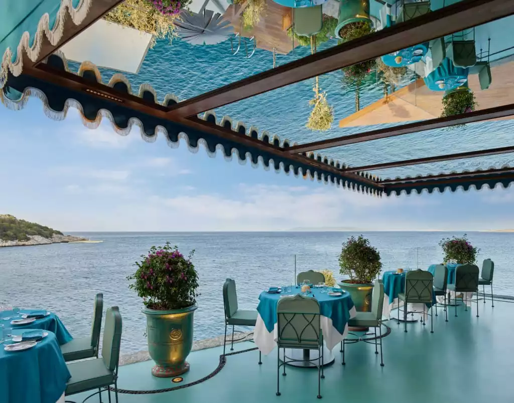 Bodrum'un muhteşem deniz manzarasına sahip bir restoran terasında, turkuaz masa örtüleri ve uyumlu sandalyelerle düzenlenmiş masalar, pembe çiçekli bitkilerle süslenmiş ve deniz yansımalarıyla bezeli ayna tavan altında huzurlu bir atmosfer sunuyor.