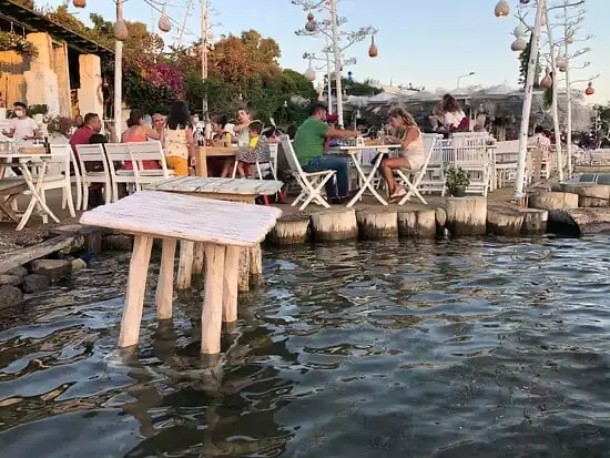 Bodrum'un eşsiz güzellikteki restoranlarından birinde, ayakları suya değen rustik beyaz ahşap masalar ve şık asılı aydınlatmalar eşliğinde misafirler, denizin kıyısında keyifli bir akşam yemeği deneyimi yaşıyor.