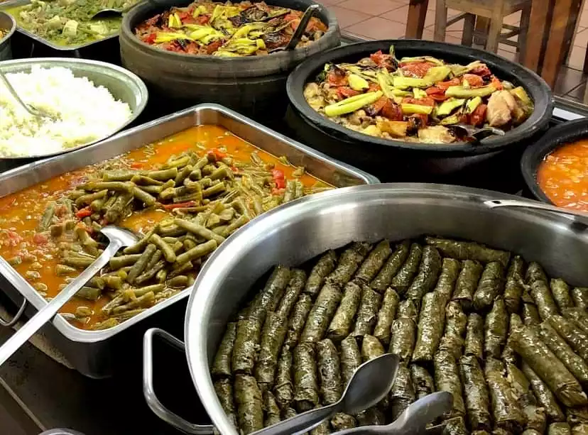 Çeşitli geleneksel Türk yemeklerinin sunulduğu zengin bir açık büfe masası: metal kaplarda domates soslu taze fasulye, pirinç pilavı ve bolca sarma, arka planda ise güveçlerde sebzeli et yemekleri gibi yöresel lezzetler dikkat çekiyor.
