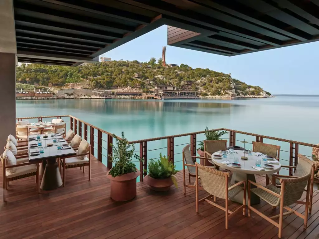 Bodrum'da, ahşap bir platform üzerinde denize nazır lüks bir restoranın geniş terasında, parlak turkuaz denizi ve karşıdaki yemyeşil yamaçların kuşatıcı manzarasıyla özenle hazırlanmış yemek masaları ve konforlu sandalyeler içeren davetkar bir atmosfer.