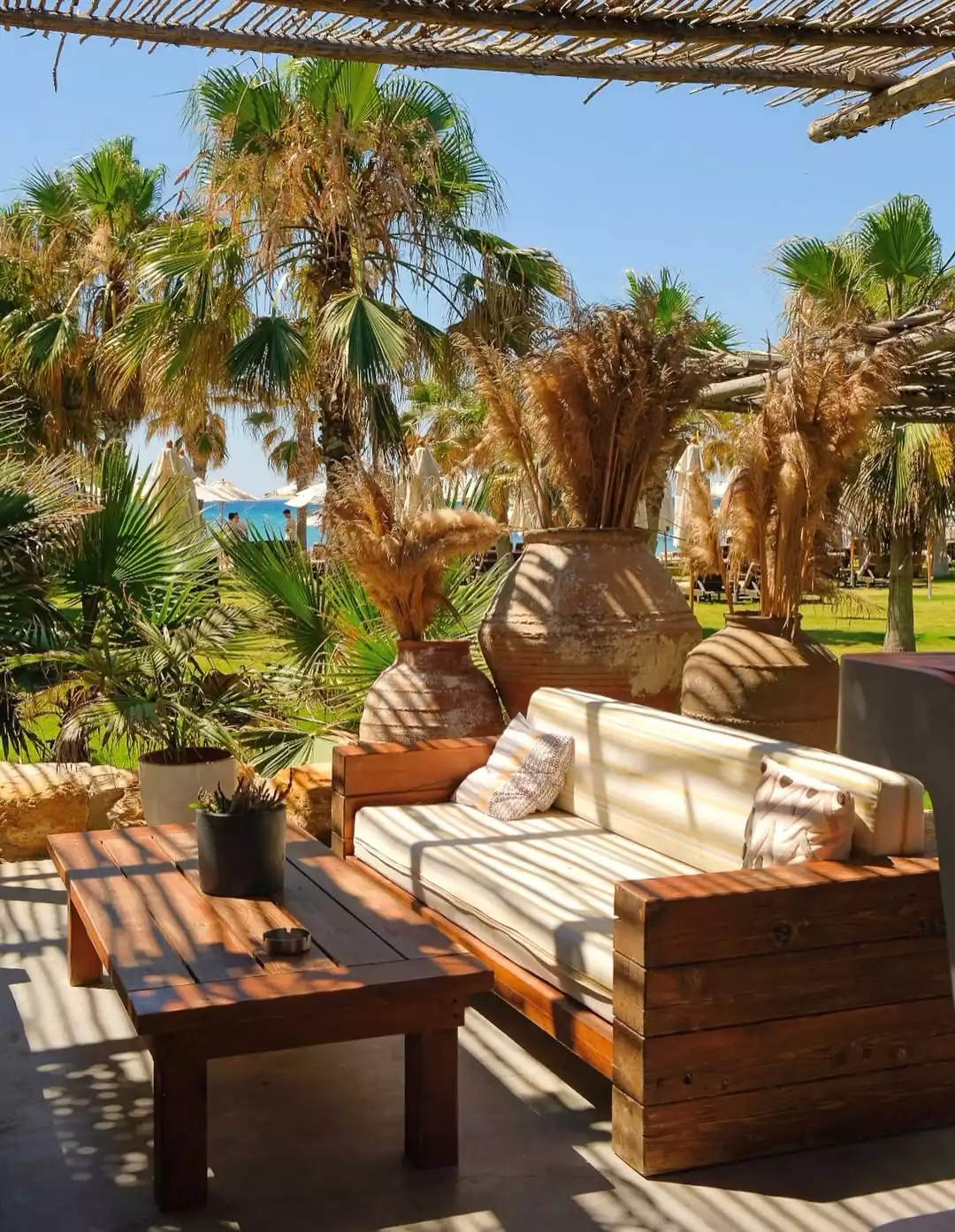 Fly-Inn Beach'teki bu dinlenme alanında, güneşin ahşap pergoladan süzülen çizgili gölgeleri altında, rahat minderli ahşap bir kanepe ve sehpa, yemyeşil palmiyeler, rustik testilerdeki kurutulmuş otlar ve uzak plaj ile denizin huzurlu manzarasıyla tropikal bir atmosfer sunuluyor.