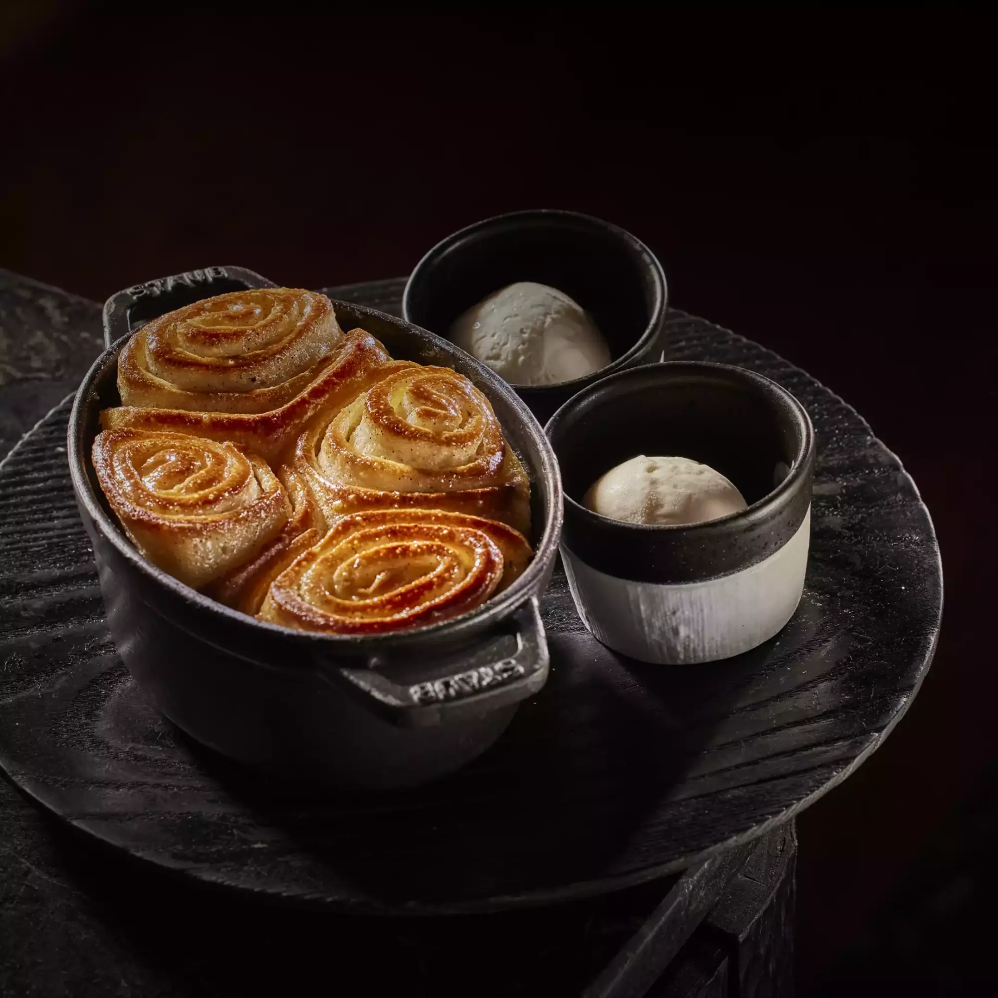 Staub marka koyu renk bir döküm kapta servis edilen, üzerleri parlayan karamelize soslu ve tarçın serpintili dört adet altın sarısı spiral çörek, yanında koyu renk seramik kaselerde iki top vanilyalı dondurma ile koyu ahşap bir tepsi üzerinde sunuluyor.