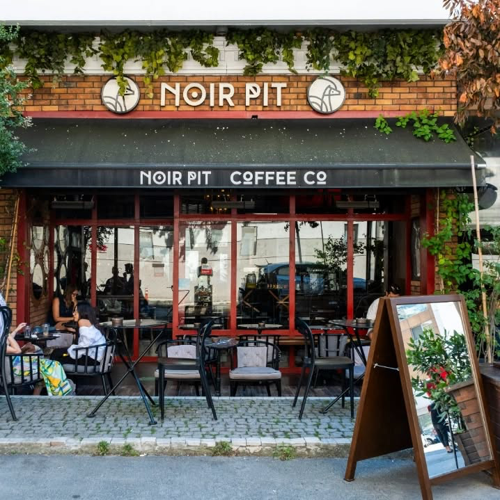 Kırmızı tuğla cepheli, siyah tente altında belirgin Noir Pit Coffee Co yazısıyla dikkat çeken, kaldırımda şık metal masalar ve sandalyelerle dolu bir açık hava kahve dükkanı önü, duvarları saran yeşil sarmaşıklar ve sağda yansıtıcı aynalı bir tabela ile canlı bir atmosfer sunuyor.