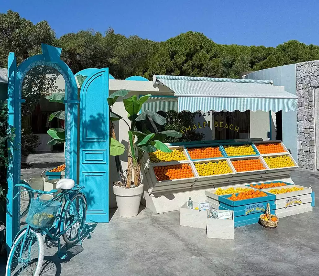 Turkuaz renkli kemerli kapı ve eski tarz bisikletin önünde, taze limon ve portakallarla hazırlanan meyve standı pastel tonlardaki sahil atmosferini tamamlıyor.