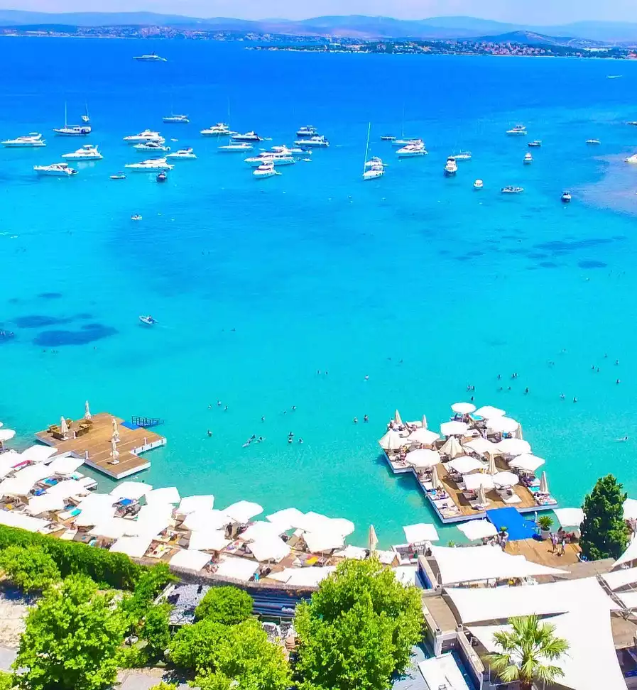Sole Mare'nin adeta cennetten bir köşe gibi görünen turkuaz renkli berrak denizinde demirlemiş çok sayıda lüks yatın bulunduğu, kıyı şeridinde ve su üzerindeki ahşap platformlarda bembeyaz şemsiyelerle donatılmış şezlonglarda tatilcilerin güneşlendiği ve serin suların tadını çıkardığı hareketli bir yaz günü plaj manzarası.