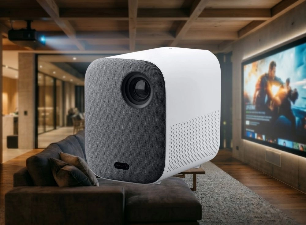 Beyaz ve gri renkli Xiaomi Mi Smart Projector 2 akıllı projeksiyon cihazı, modern bir oturma odasının şık atmosferinde, arkasında büyük bir ekranda aksiyon filmi oynarken koyu renkli bir kanepenin önünde duruyor. Bu 1080p full HD projeksiyon, Android TV ve Google Asistan özellikleriyle gelişmiş bir ev sineması deneyimi vadediyor.