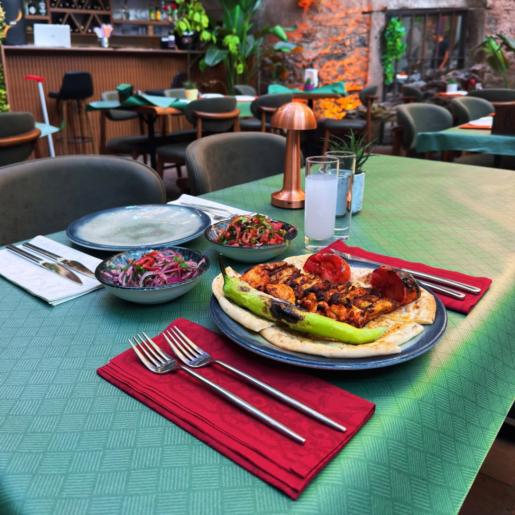 Arka planda yeşilliklerle dolu modern bir restoranın görüldüğü samimi bir ortamda, desenli yeşil masa örtüsü serili masada servis edilmiş ızgara tavuk, közlenmiş biber, domates ve iki çeşit taze salatanın yanı sıra rakı ve su bardakları bulunuyor.