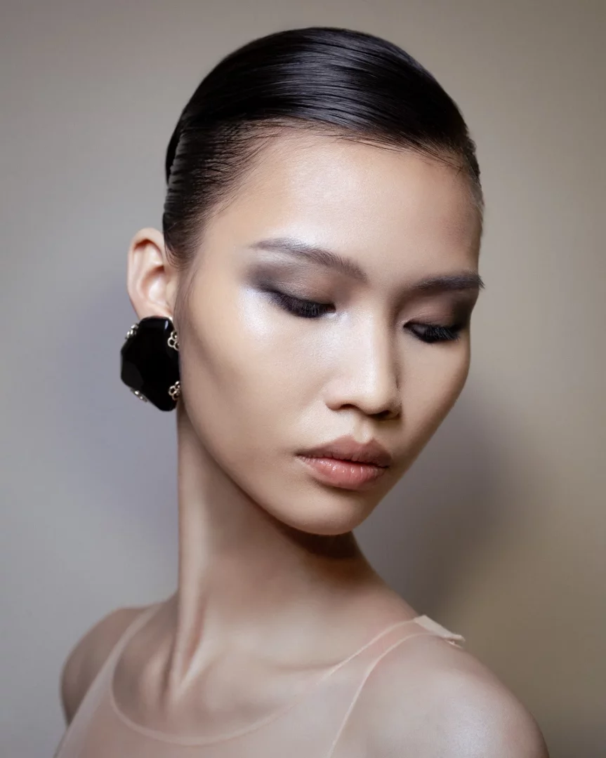 Bir modelin aşağı bakıp hafifçe kapalı gözlerle durduğu, Armani sonbahar-kış güzellik görünümünü sergileyen bu yakın çekimde, ıslak görünümlü toplanmış saçları, dumanlı gri farlarla vurgulanmış gözleri, ışıltılı teni ve büyük siyah küpesi dikkat çekiyor.