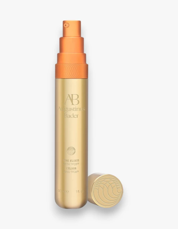 Beyaz arka planda duran, altın rengi Augustinus Bader The Elixir serum şişesi, turuncu pompalı üst kısmı ve yanında duran desenli altın kapağıyla ürünün lüks cilt bakımı özelliklerini vurguluyor.