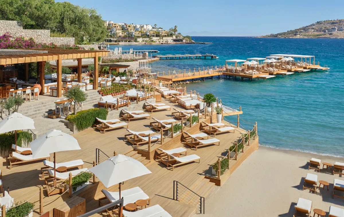 The Bodrum EDITION beach club alanında beyaz kum plaj, ahşap iskeleler, şezlonglar ve Yalıkavak deniz manzarası