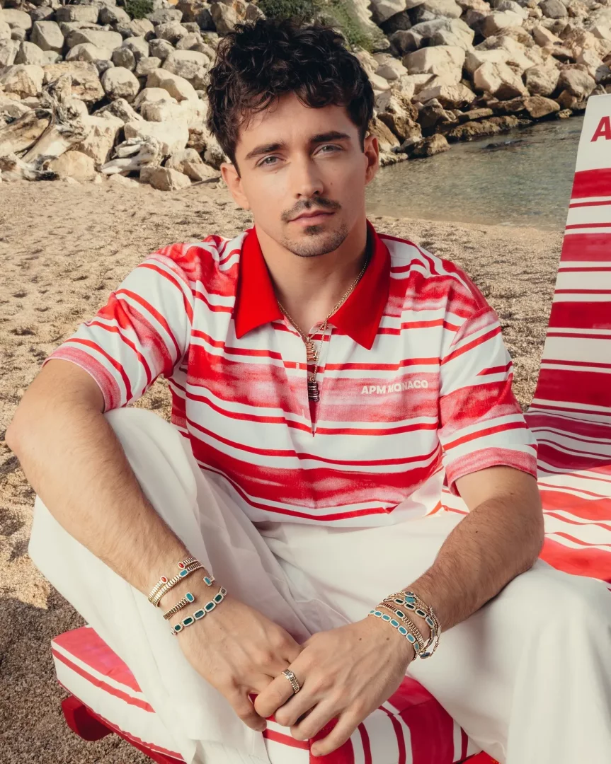 Charles Leclerc, kırmızı beyaz çizgili polo tişörtü ve beyaz pantolonuyla kumsalda kırmızı beyaz çizgili bir şezlongda oturmuş, kolundaki şık bileklikler ve yüzüklerle APM Monaco markasının zarafetini vurgularken doğrudan objektife bakıyor.