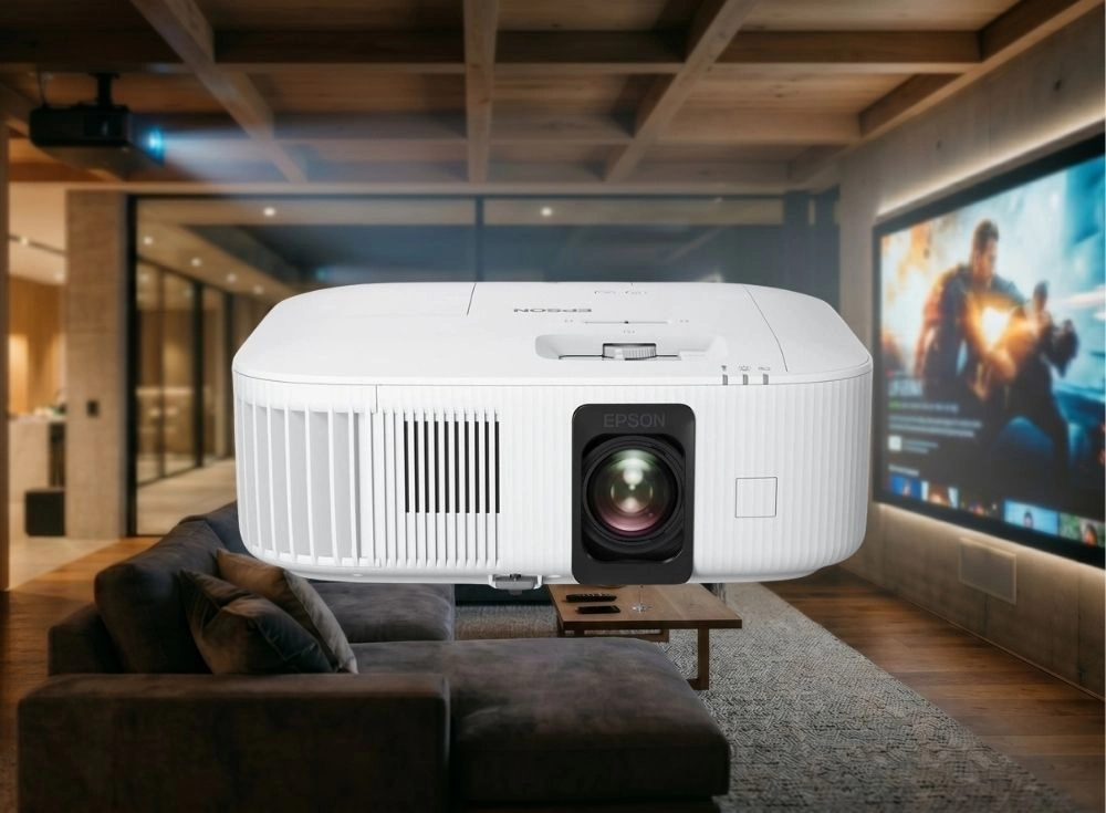 Modern ve sıcak bir ev sineması ortamında, koyu gri rahat bir L koltuğun ve ahşap orta sehpanın önünde belirgin bir şekilde havada süzülür gibi duran beyaz renkli bir Epson akıllı projeksiyon cihazı, arka planda büyük bir ekranda gösterilen heyecan verici bir aksiyon filmi sahnesi ile uyum içinde.