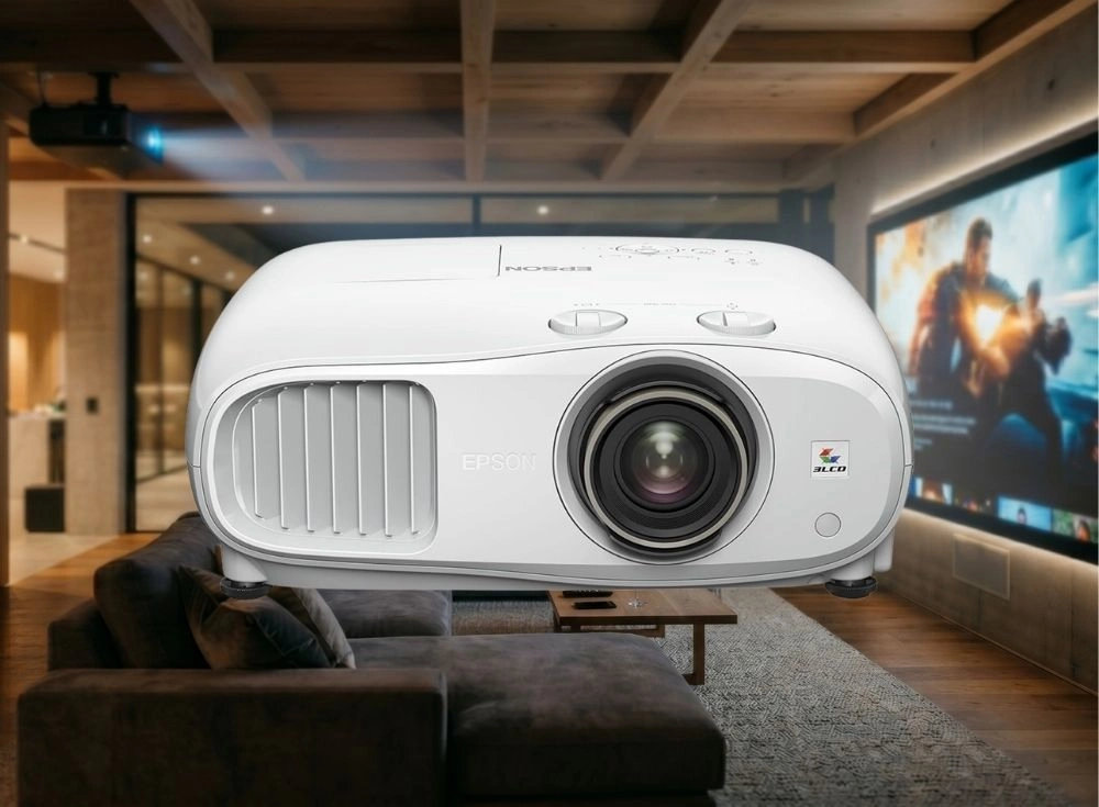 Beyaz renkli, ön kısmında büyük bir lens ve havalandırma ızgarası olan Epson EH-TW7100 4K Pro-UHD ev sineması projeksiyon cihazı, tavanında başka bir projeksiyon cihazının da bulunduğu, geniş bir ekranda aksiyon filmi gösterilen ve koyu renkli koltukların yer aldığı modern bir ev sinema ortamında öne çıkıyor.