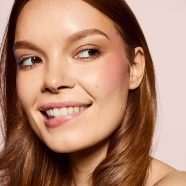 Fenty Beauty Cheeks Out Freestyle Krem Allık ürününün doğal ve ışıltılı etkisini vurgulayan bu yakın çekimde, hafifçe dudaklarını ısırırken gülümseyen genç bir kadının yanaklarında pembemsi ve taze bir allık tonu fark ediliyor.