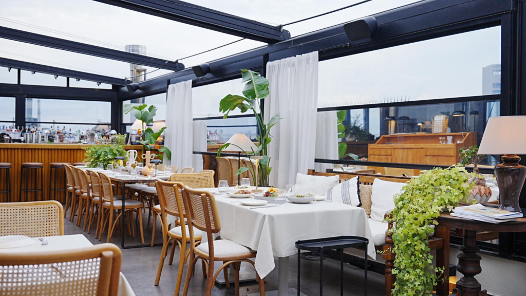 Bu aydınlık çatı katı restoranında, uzun bir masanın etrafında düzenlenmiş hasır sandalyeler, beyaz masa örtüleri ve üzerlerinde çeşitli yemekler bulunan tabaklar seçiliyor; arka planda içkilerin sıralandığı şık bir bar, büyük yeşil bitkiler ve şehir silüetine bakan geniş cam pencereler mekanı ferah bir atmosfere kavuşturuyor.