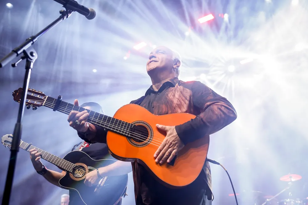 Sahne ışıkları ve sahne sisi arasında, turuncu klasik gitarını çalan, başını hafifçe yukarı kaldırmış, yüzünde mutlu bir ifadeyle coşkulu bir performans sergileyen yaşlı bir müzisyen ve arkasında beliren diğer gitarist, enerjik bir konser anını yansıtıyor.