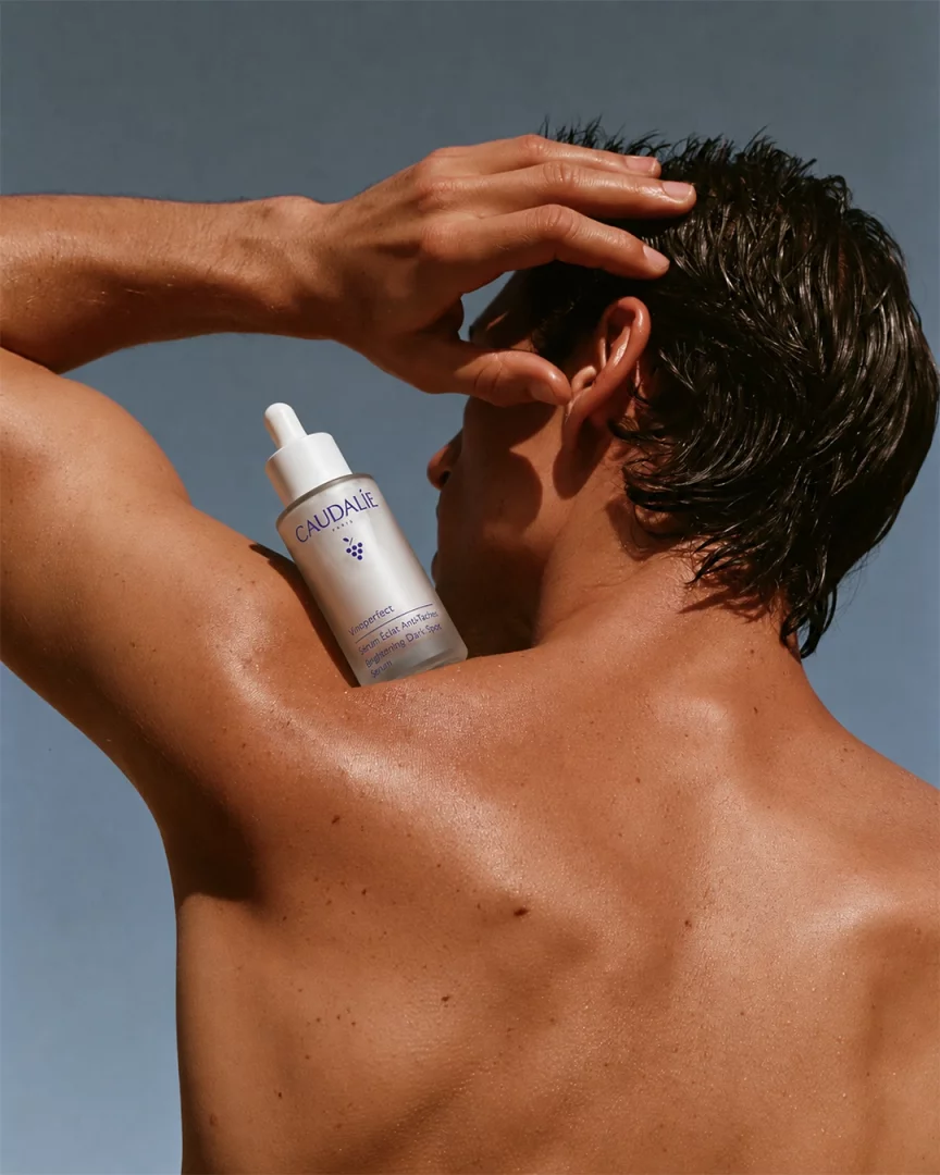Bronzlaşmış teni ve ıslak saçlarıyla görünen bir erkeğin omzunda duran Caudalie Vinoperfect Sérum Eclat Anti-Taches leke karşıtı aydınlatıcı serum şişesi, cilt bakımında güneşin etkilerine karşı etkili bir bakımı ve leke giderme önerisini sunuyor.