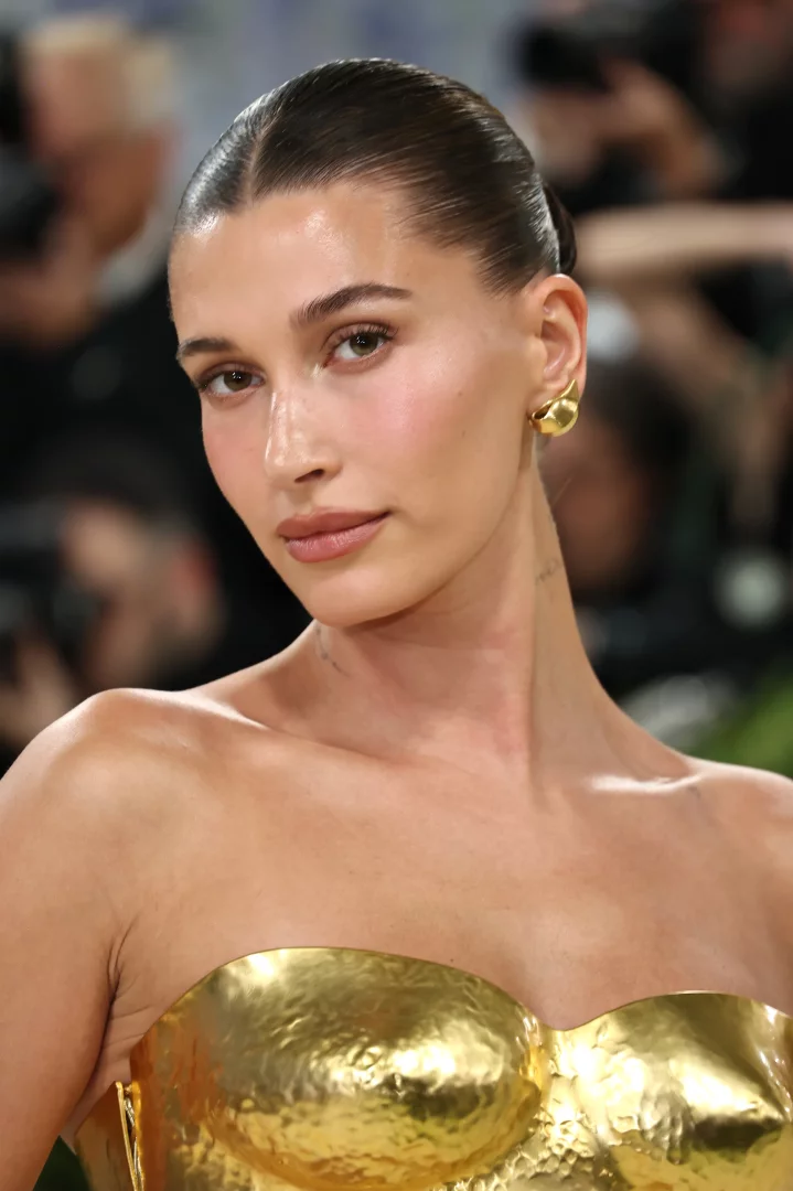 Hailey Bieber'ın çekici bir ifadeyle kameraya baktığı yakın çekim portresinde, arkaya taranmış saçları, doğal parlak cildi ve altın rengi metalik straplez kıyafeti ön plana çıkarken, kahverengi göz makyajı dikkat çekiyor.
